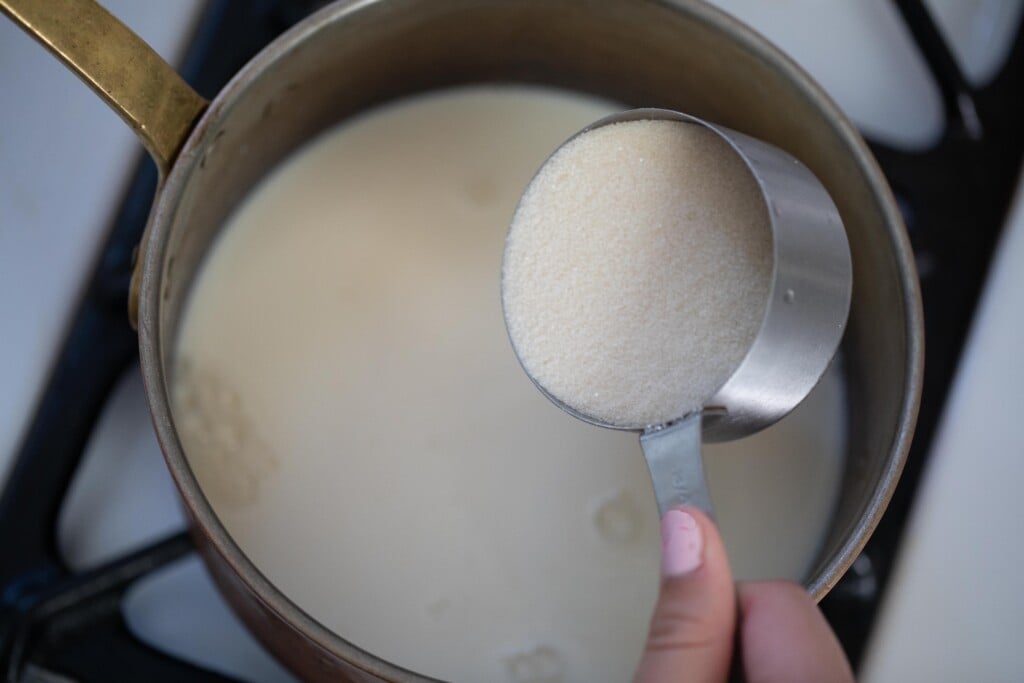 A hand adding a measuring cup full of sugar to a saucepan of milk.