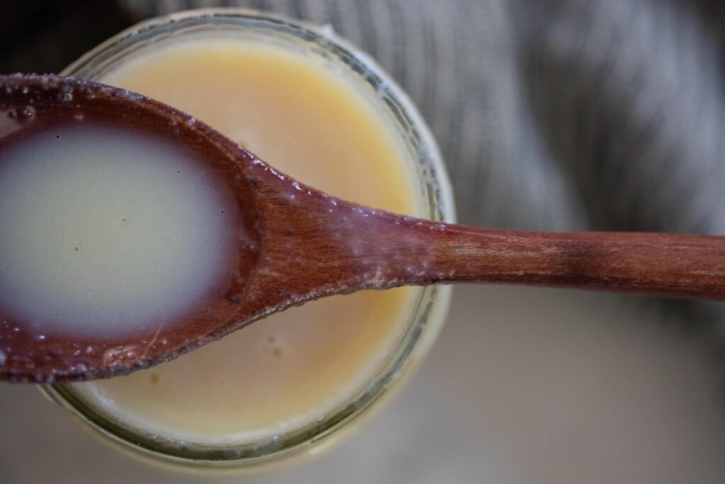 A wooden spoon laying over the top of an opened jar full of sweetened condensed milk.