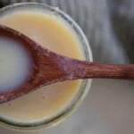 A wooden spoon laying over the top of an opened jar full of sweetened condensed milk.