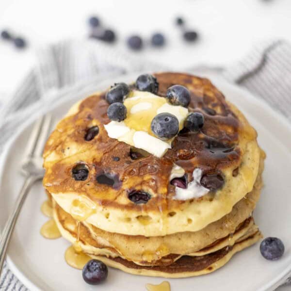 a stack of sourdough blueberry pancakes with a pat of butter and honey on top. The pancakes are on a white plate.