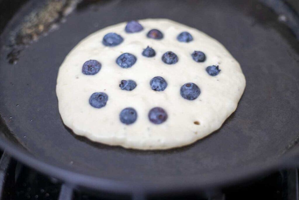 a pancake with blueberries frying on a cast iron skillet.