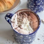 A blue mug of hot chocolate with whipped cream and chocolate shavings on top next to a plate of croissants.