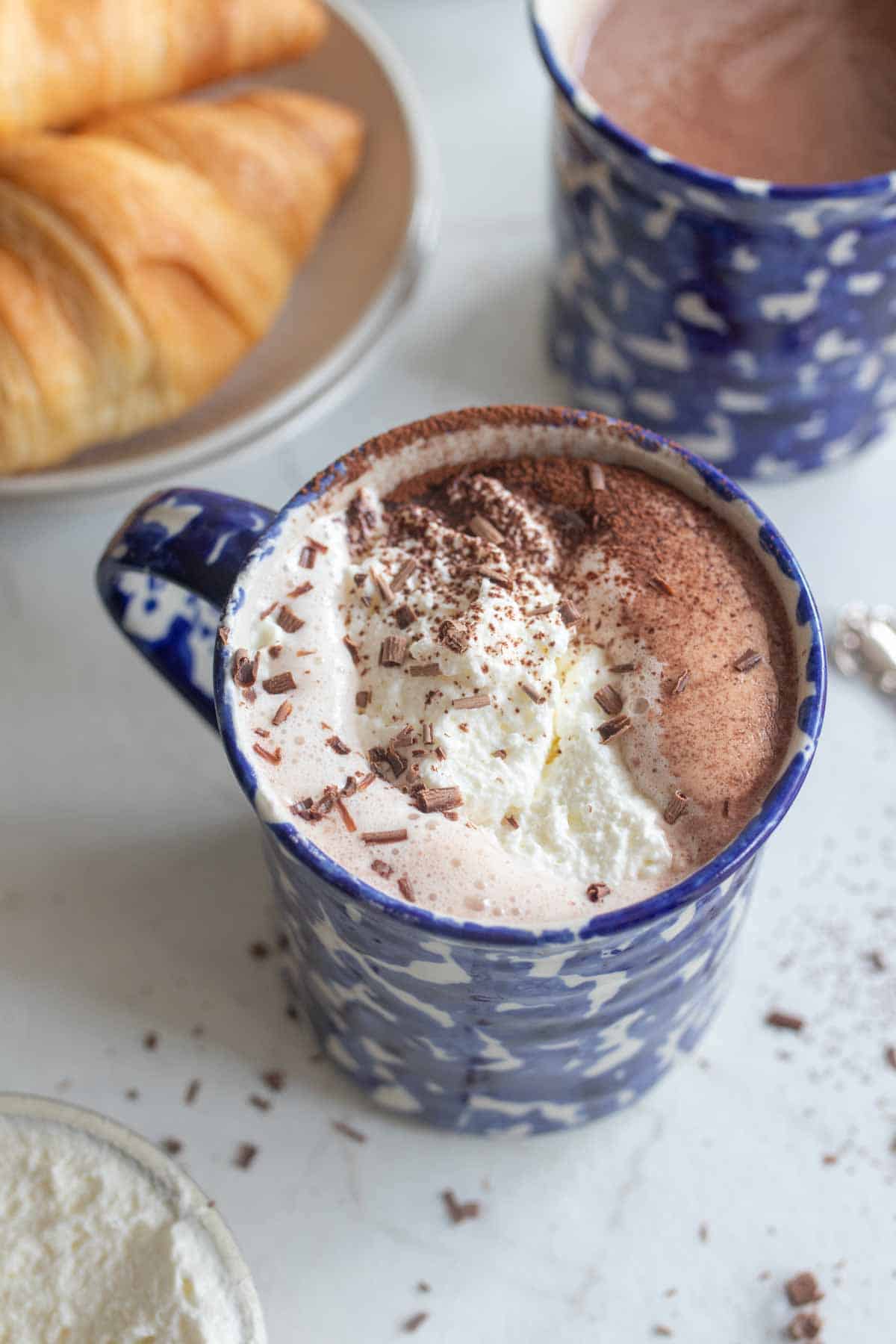 A blue mug of hot chocolate with whipped cream and chocolate shavings on top next to a plate of croissants. 