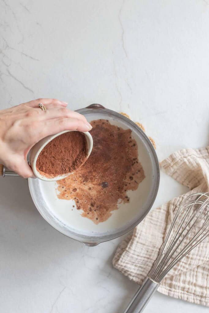 Someone pouring cocoa powder into a saucepan of milk. 