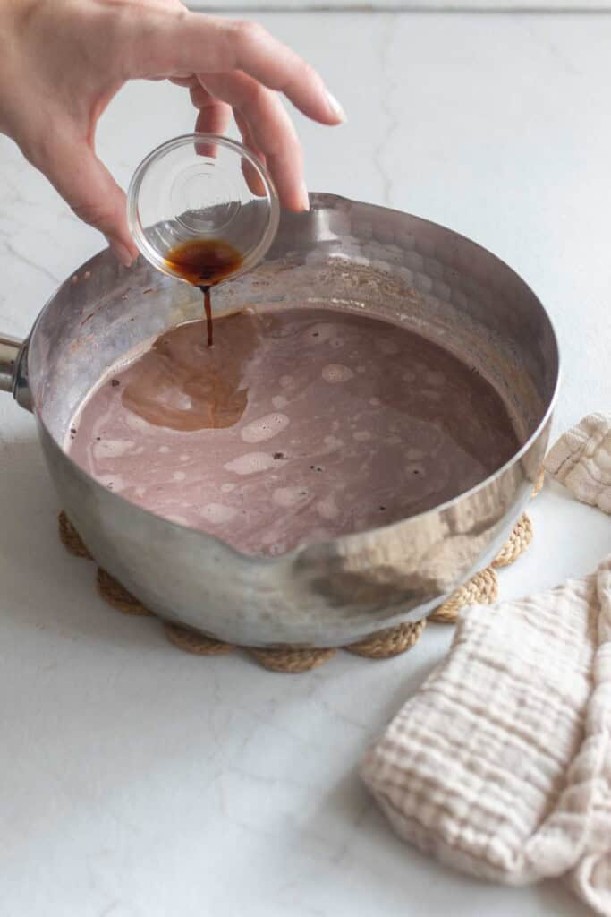 A hand pouring in vanilla to a saucepan of chocolate milk.