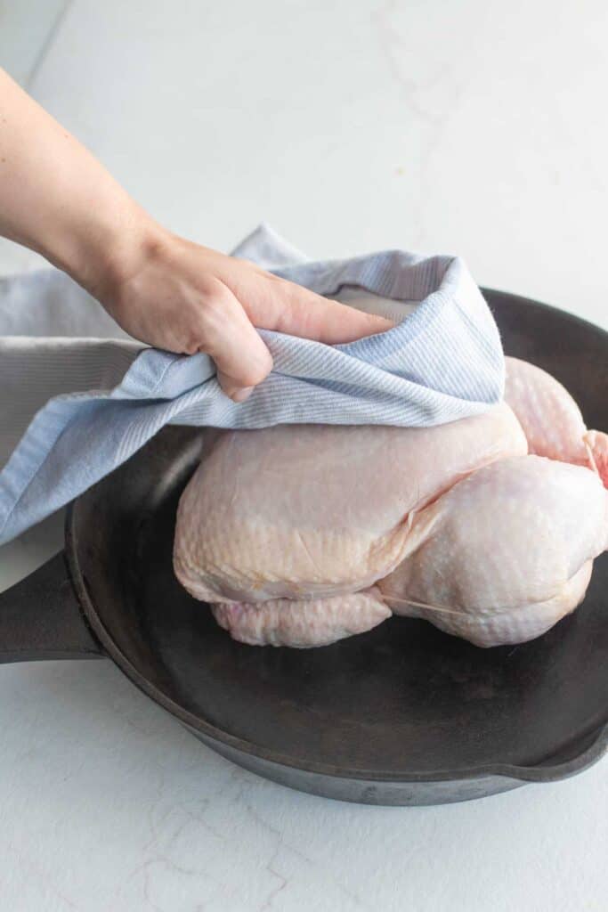 A whole chicken in a cast iron skillet with a woman's hand patting it dry with a blue hand towel.