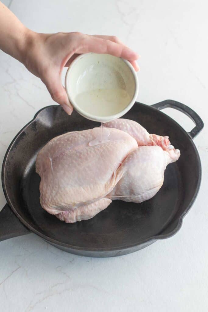 A hand drizzling oil from a white container onto a whole chicken in a cast iron skillet.