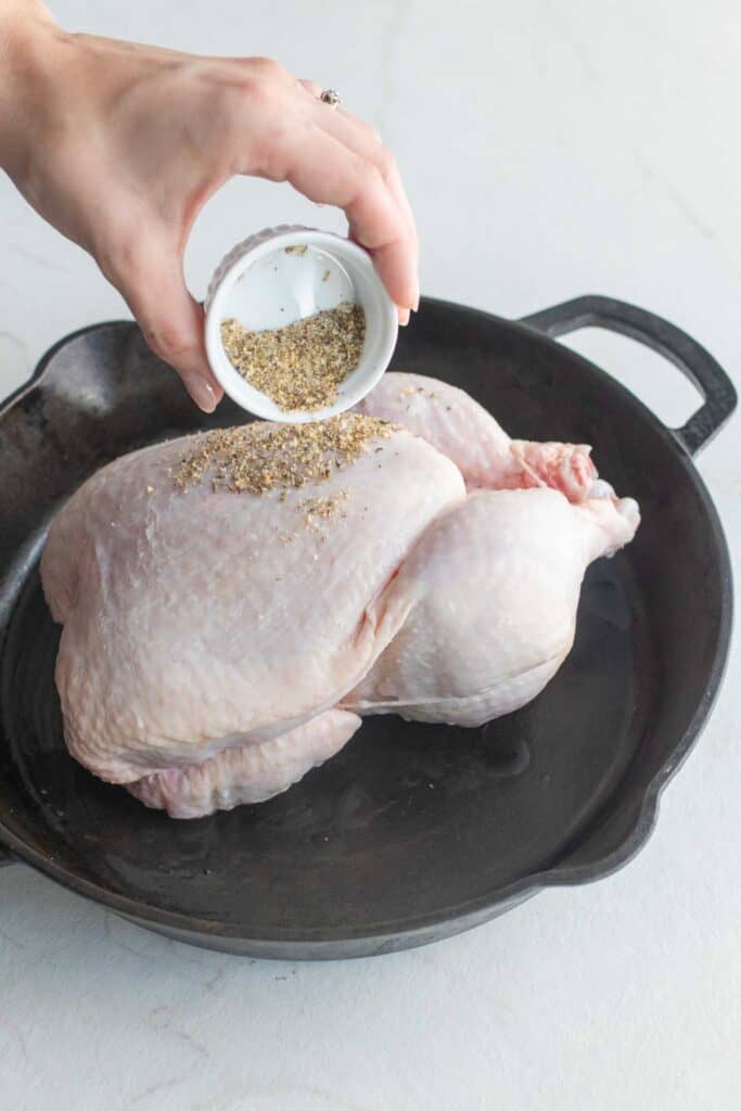 A hand sprinkling seasoning from a white dish onto a whole chicken in a cast iron skillet.