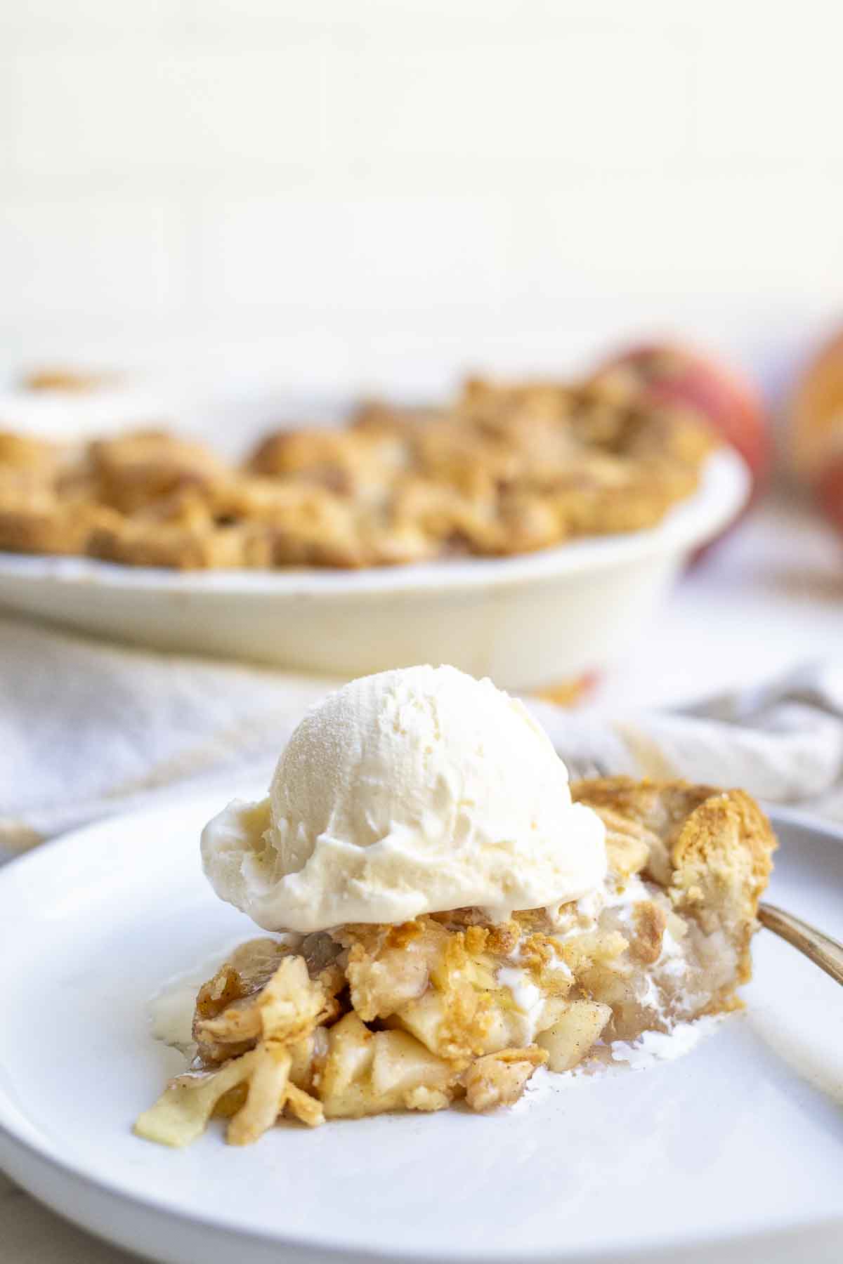 slice of apple pie topped with a scoop of ice cream on a white plate. More pie is in the background.