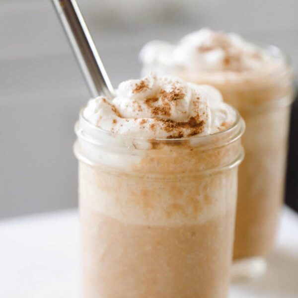 two pumpkin smoothies topped with whipped cream sprinkled with pumpkin spice. A metal straw is in the smoothie and the smoothies sit on a white countertop.