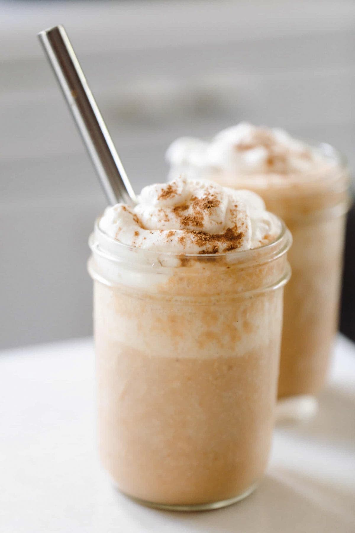 two pumpkin smoothies topped with whipped cream sprinkled with pumpkin spice. A metal straw is in the smoothie and the smoothies sit on a white countertop.