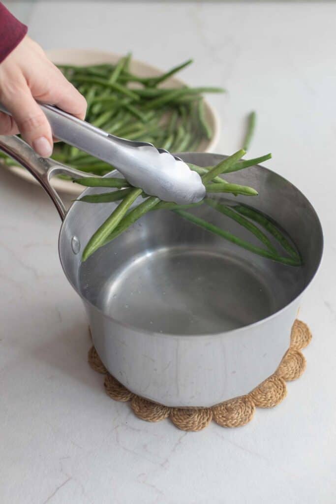 A hand holding tongs filled with green beans and adding them to a pot of water.