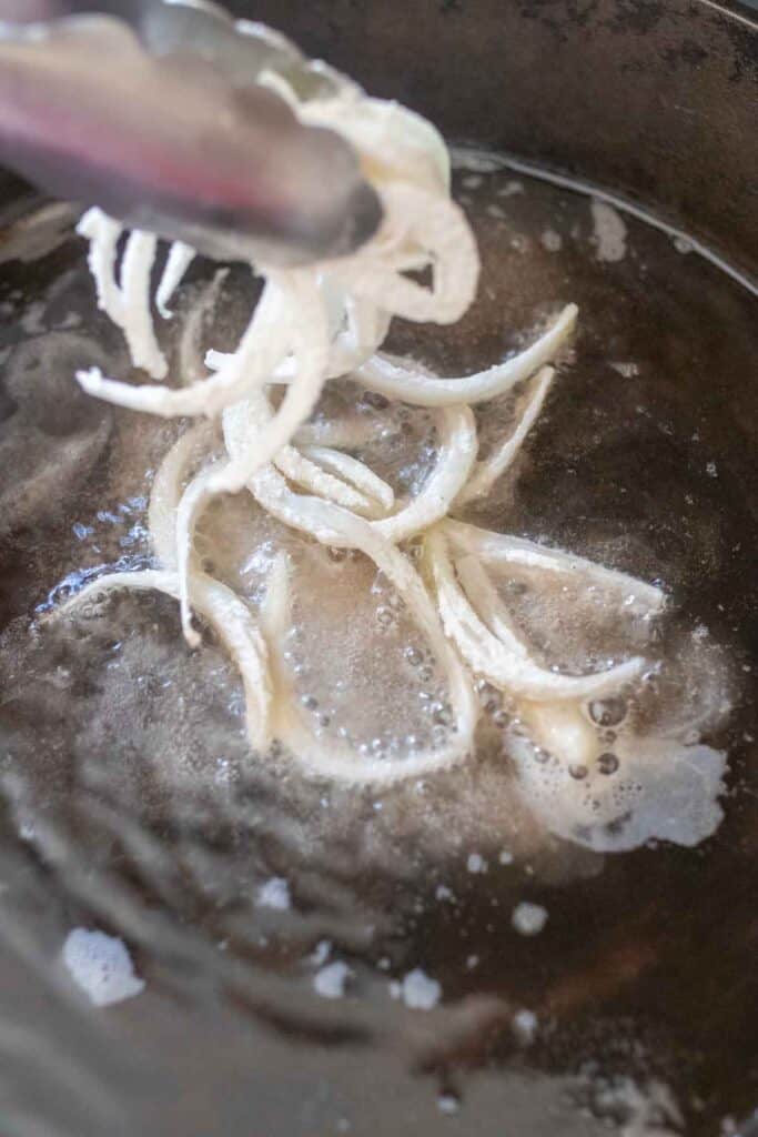 Tongs throwing in seasoned sliced onions into a heated cast iron skillet.