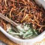 A casserole dish filled with green bean casserole with a wooden spoon scooping out a serving.