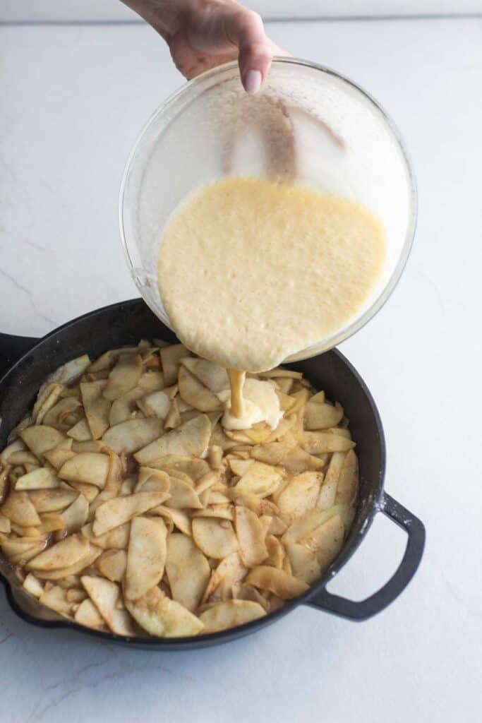 Someone pouring the sourdough apple cobbler topping that is in a glass bowl over the sliced cooked apples in a cast iron skillet.