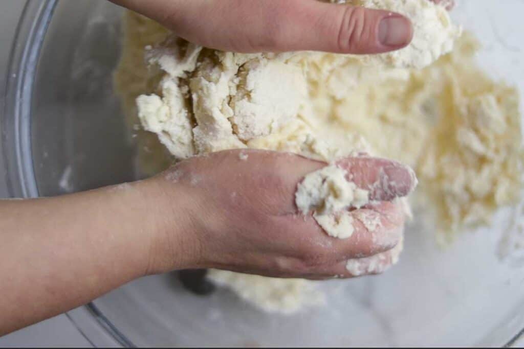 Two hands working pie dough to help it come together in a bowl. 