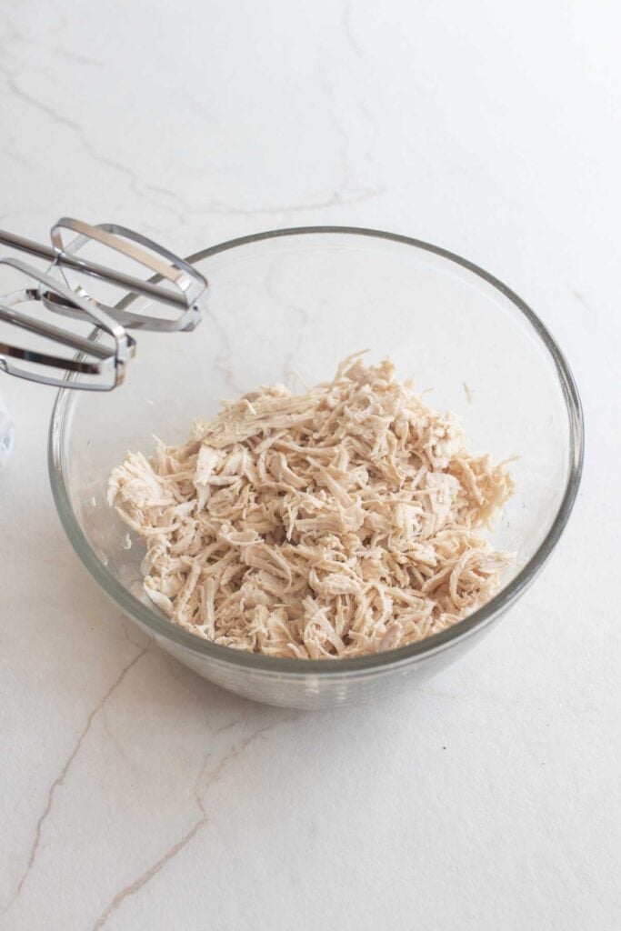 Shredded chicken in a small bowl with a hand mixer in the background.