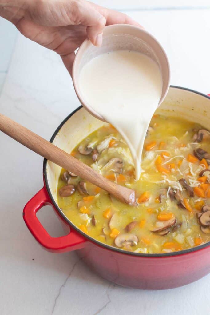 Heavy cream poured into a dutch oven full of chicken marsala soup with a wooden spoon sticking out.