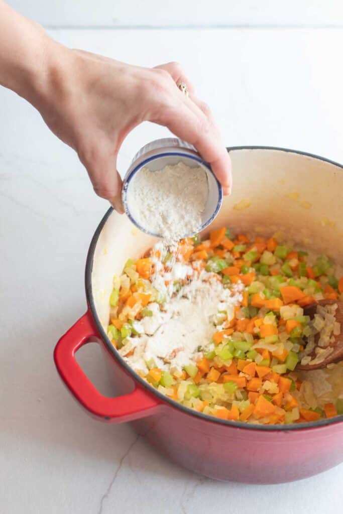 A hand adding a little flour to a dutch oven of sauted veggies.