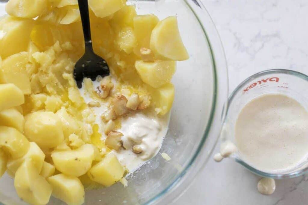 Potatoes, roasted garlic, butter and cream in a glass bowl. A measuring cup of milk sits to the side.