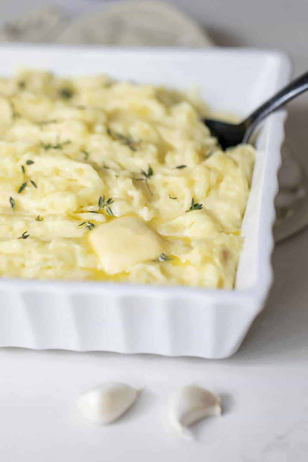 White baking dish with garlic mashed potatoes topped with herbs with a spoon in the potatoes.