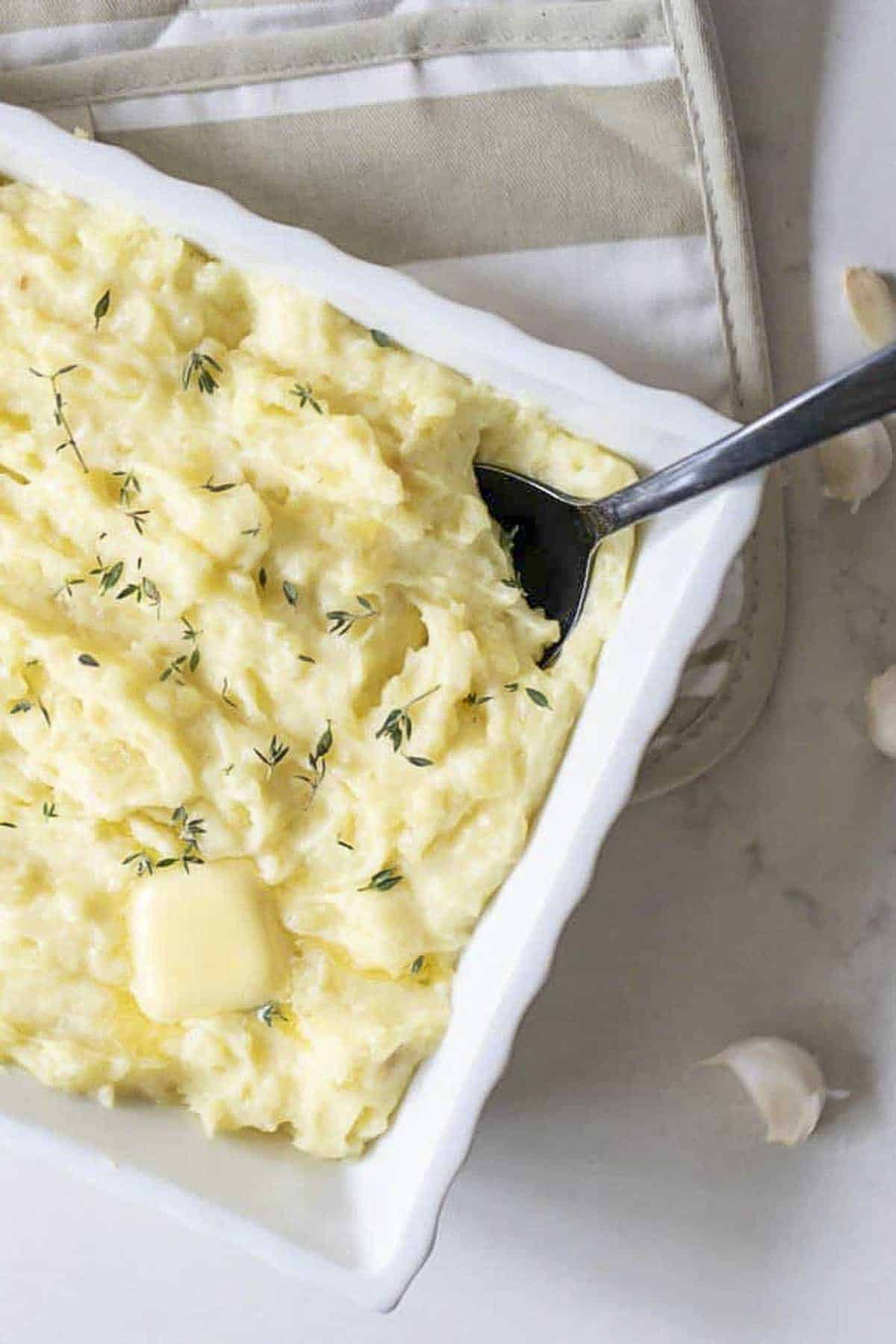 Photo of garlic mashed potatoes with herbs sprinkled on top in a white baking dish on a tan stripped pot holder.