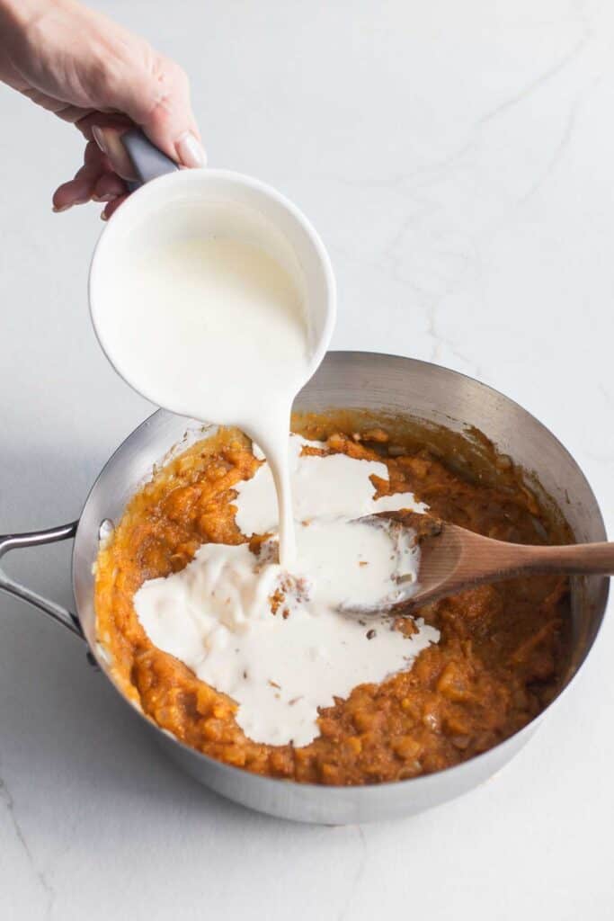 Heavy cream poured from a dish into a stainless steel pot full of pumpkin pasta sauce.