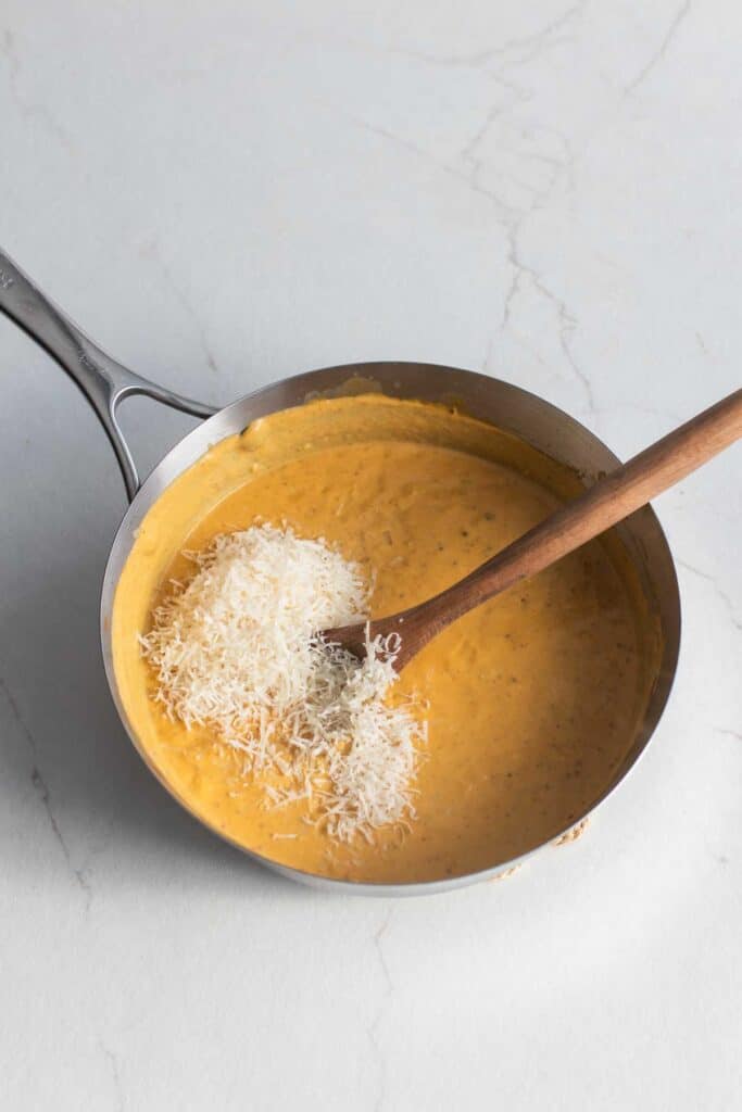 Grated cheese added to a stainless steel pan of sauce with a wooden spoon.