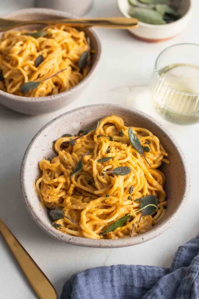 Two bowls full of spaghetti noodles covered in pumpkin pasta sauce topped with fried sage leaves.