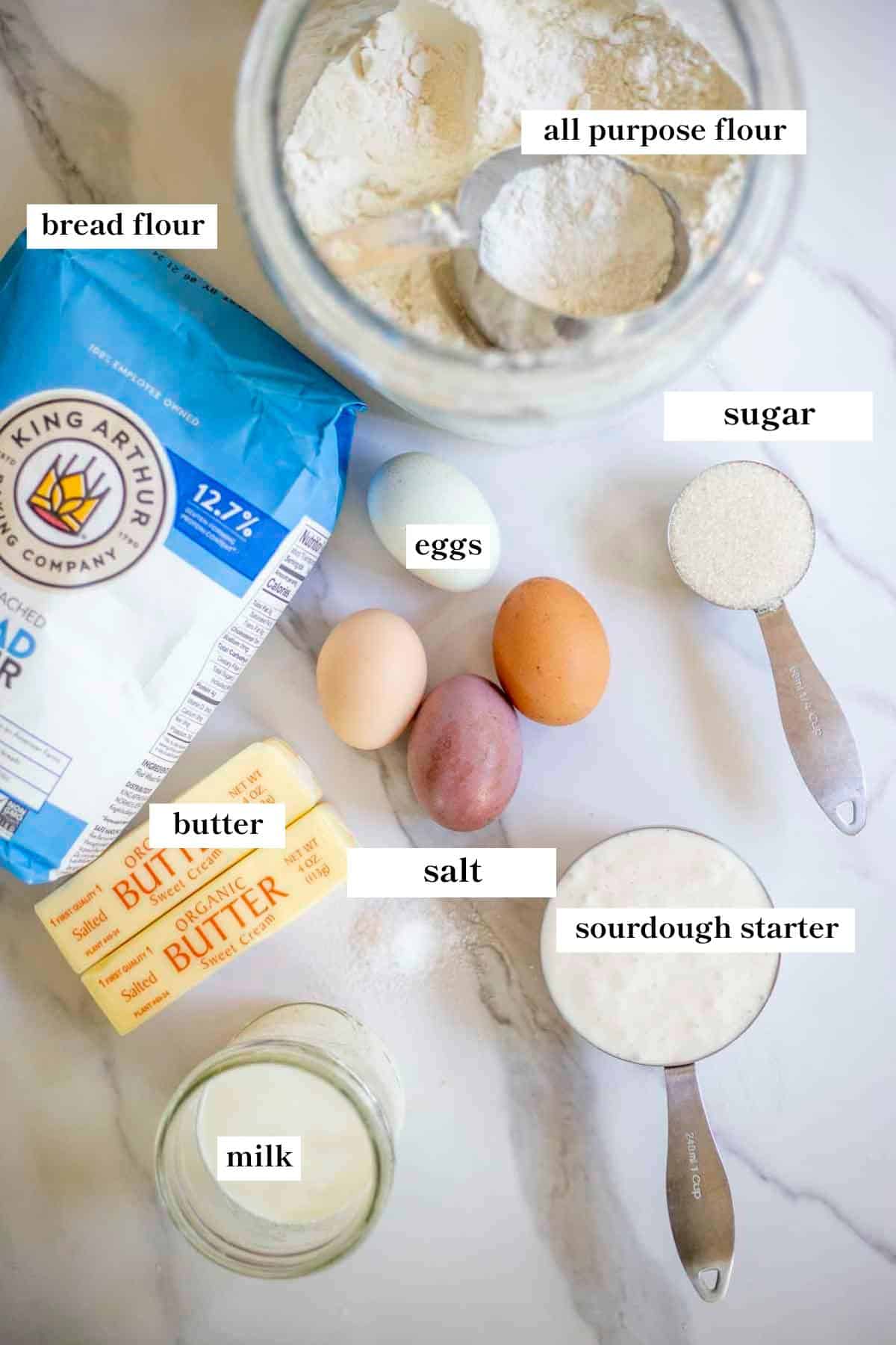 Flour, butter, eggs, sugar, salt, sourdough starter, and milk on a countertop.