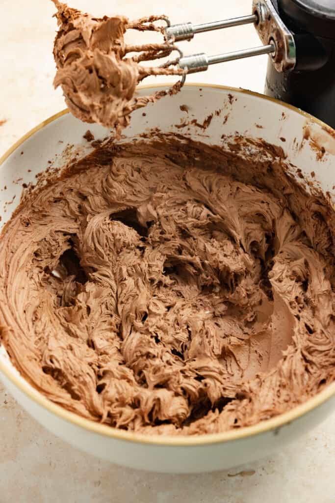 Chocolate frosting mixed in a bowl with a hand mixer next to it.