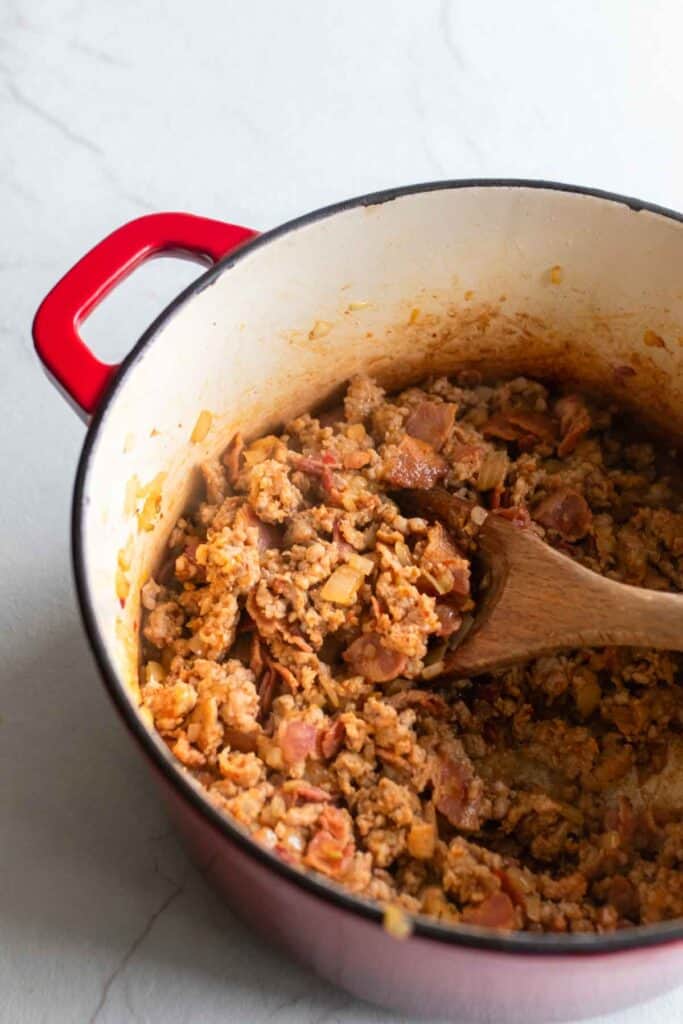 Sausage, onions, and bacon cooking in a dutch oven with a wooden spoon stirring.