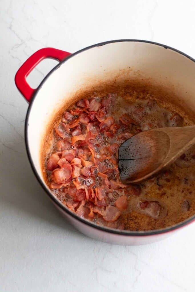 A dutch oven with bacon simmering with a wooden spoon sticking out. 