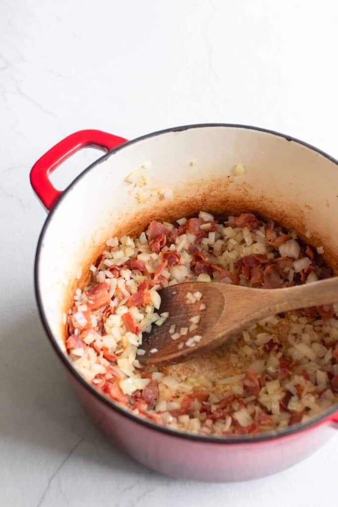 A dutch oven with bacon and onions with a wooden spoon stirring.