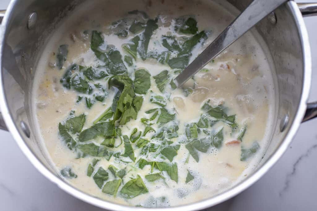 Spinach and cream added to chicken and gnocchi soup in a large stock pot with a spoon.