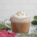 Peppermint mocha topped with whipped cream and sprinkles in a glass mug. The coffee drink is surrounded by fresh greenery.