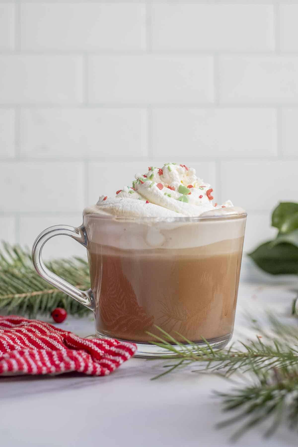 Peppermint mocha topped with whipped cream and sprinkles in a glass mug. The coffee drink is surrounded by fresh greenery.