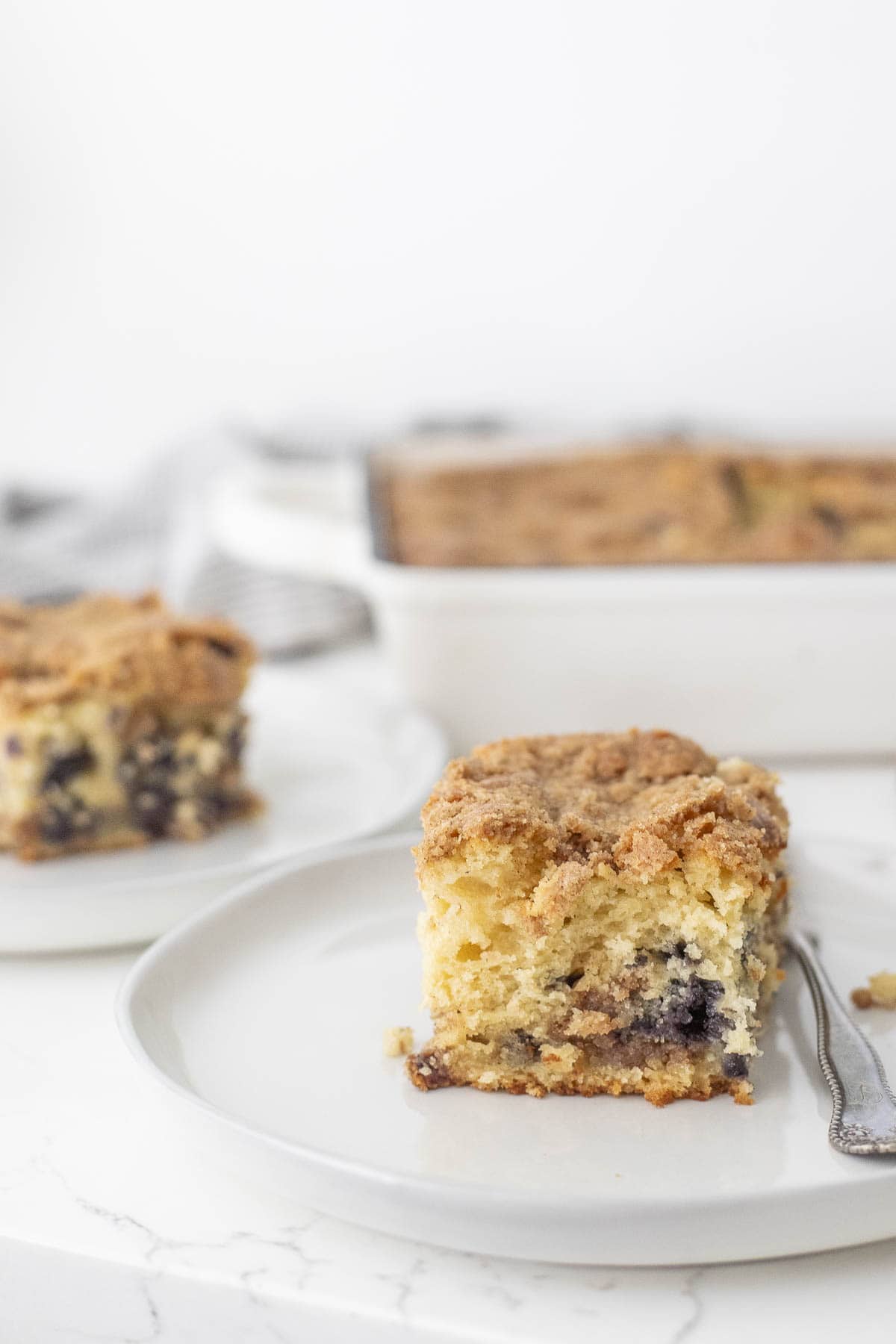 a slice of sourdough blueberry coffee cake on a plate with more coffee cake in the background.