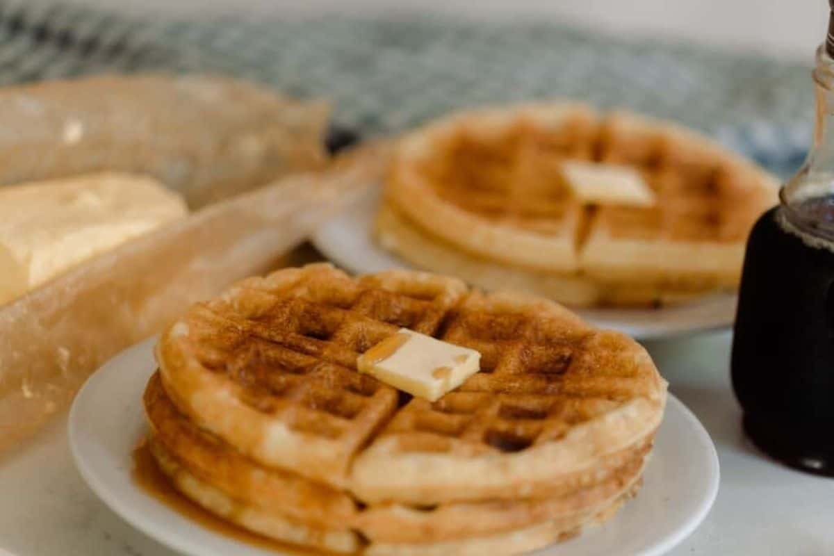 Two stacks of waffles on a small plates with a slab of butter and maple syrup on top.