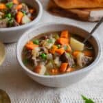 A bowl of beef stew with a loaf of bread in the background.