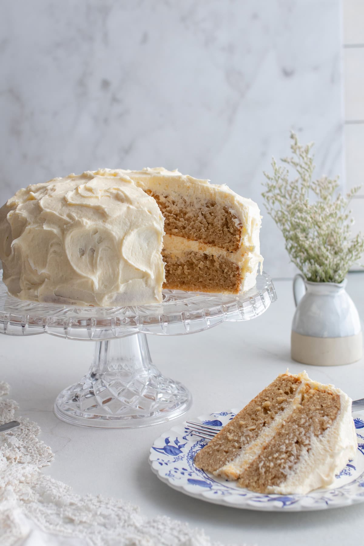 Earl grey cake with cream cheese frosting on a glass cake stand with a slice taken out and on a blue and white plate.