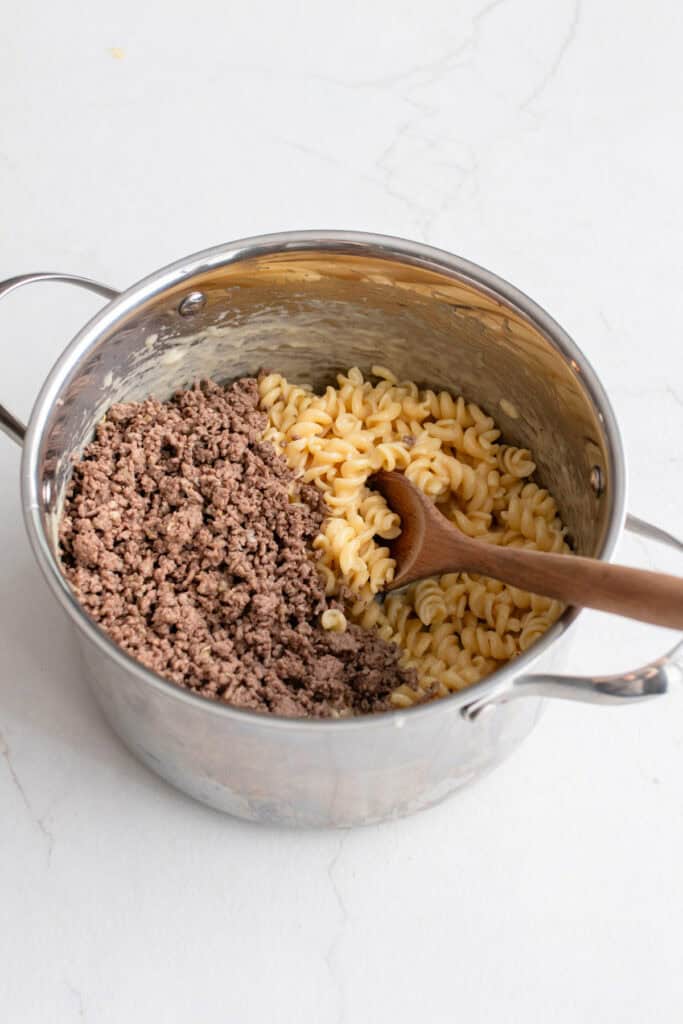 Browned beef added to a pot of pasta with a wooden spoon.