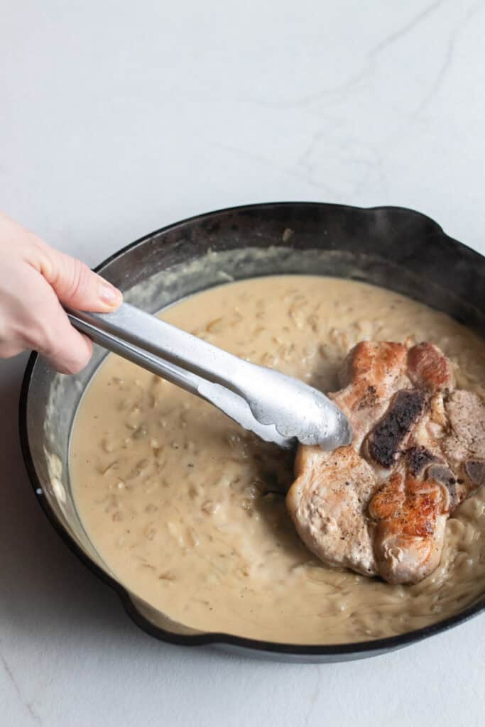 Adding pork steaks back into the cast iron skillet with creamy onion sauce.