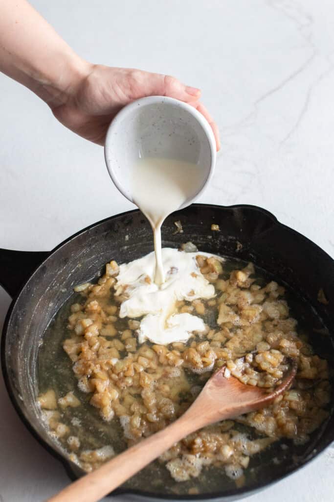 Adding cream to sautéed garlic, onions, broth, and other ingredients in a cast iron skillet.