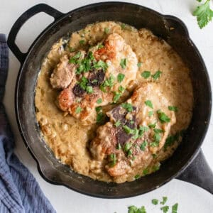 Overhead photo of pork steaks topped with fresh chopped parsley in a creamy onion gravy in a cast iron skillet. More parsley surrounds the skillet.