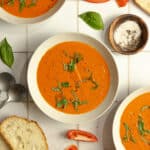 Three bowls of roasted red pepper and tomato soup topped with fresh basil.