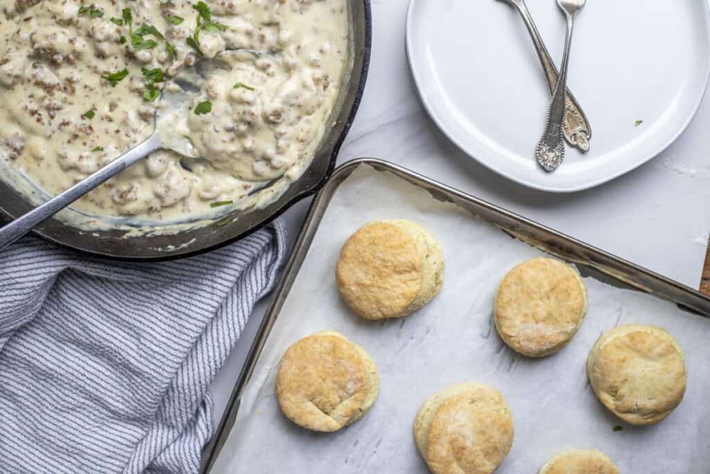 A cast iron skillet full of gravy next to a sheet pan of freshly baked biscuits.