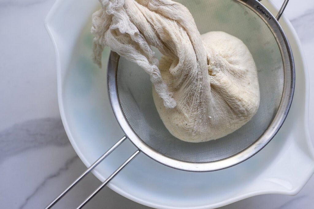 Cheese in cheesecloth with the remaining whey being squeezed out over a bowl.