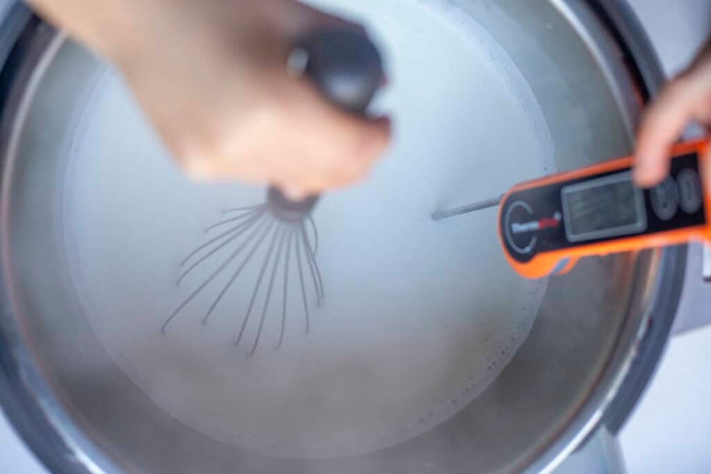 Whisking milk with one hand and holding a thermometer in the milk in the other hand.