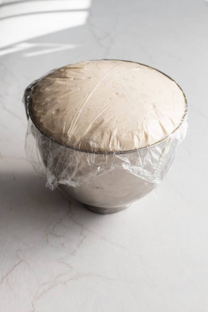 Dough in a stand mixer bowl risen to the top and covered in plastic wrap.